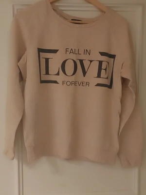 Beige sweatshirt från Amisu med tryck - Säljer en beige sweatshirt från Amisu i storlek M med svart texttryck 'Fall in Love Forever' på framsidan. Tröjan har rund hals, långa ärmar och är gjord i mjuk bomullsmix. Perfekt för chill dagar och enkel att matcha med jeans eller leggings.