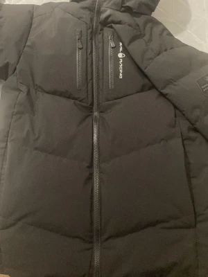 Svart pufferjacka från Sail Racing - Svart pufferjacka från Sail Racing med huva och snygga logodetaljer på bröstet och ärmen. Jackan har två vertikala bröstfickor med dragkedja och en stor patch med 'Sail Antarctica Expedition'. Perfekt för kalla dagar och har en clean, sportig look. !!PRISS KAN DISKUTERAS!!!