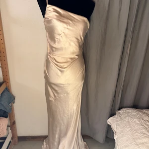 Champagnefärgad långklänning Boohoo - Säljer en elegant långklänning från Boohoo i champagnefärgad satin med smala axelband och figurnära passform. Klänningen har en slät yta, lätt glans och dragkedja bak. Perfekt för dig som vill ha en stilren och lyxig look.