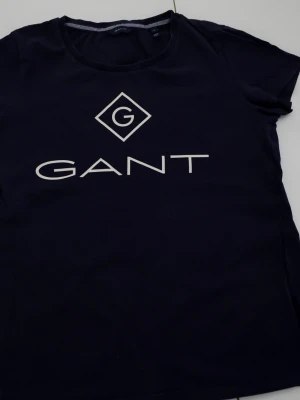 Mörkblå GANT t-shirt med logga - Snygg mörkblå t-shirt från GANT med stor vit logga på bröstet. Klassisk rund hals och korta ärmar. Tillverkad i mjuk bomull som känns skön mot huden. Perfekt för dig som gillar stilrena och enkla plagg med tydlig branding.