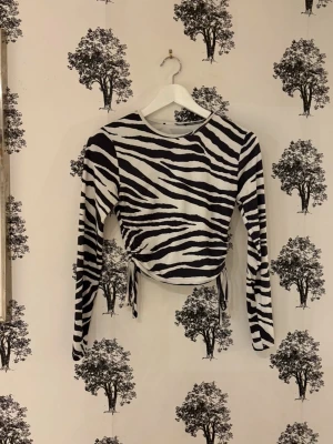 Zebramönstrad långärmad topp - Säljer en långärmad topp med svartvitt zebramönster och snörning i sidorna. Modellen är croppad och har rund hals. Materialet känns mjukt och stretchigt, troligen polyester eller bomullsmix. Perfekt för dig som vill sticka ut med ett djurmönster.