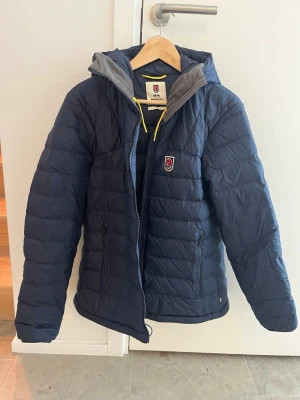 Blå Fjällräven Expedition tunnare dunjacka L - Blå Fjällräven, tunnare dunjacka i storlek L. Jackan har huva, dragkedja och klassisk Fjällräven-logga på bröstet med gul dragsko. Använd en säsong. Nypris 4499 sek