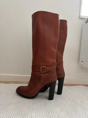 Bruna höga stövlar med klack - Snygga bruna stövlar i skinn med hög klack. Stövlarna har en dekorativ rem med spänne i mässing och dragkedja på insidan för enkel påtagning. Perfekta.Ett par bruna stövlar med hög klack..  I mycket gott skick, endast använda en gång men inser att de är för stora för mig.  Mått i cm:  Innersula: 25,5 Klack: 11,5 Total höjd: 48 Bredden toppen på skaftet: 18-19