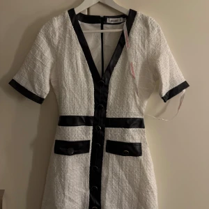 Vit kortklänning från Missguided - Säljer en vit kortklänning från Missguided med svarta detaljer och knappar framtill. Klänningen har v-ringad hals, korta ärmar och två fickor med dekorativa knappar. Materialet är strukturerat och ger en lyxig känsla. Perfekt för dig som gillar kontraster och stilrena plagg.