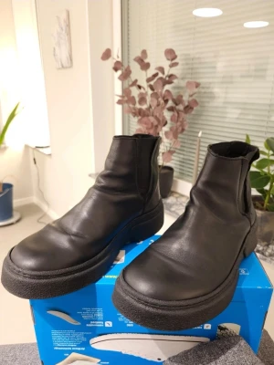 Svarta Björn Borg chelsea boots i skinn 42 - Säljer ett par svarta chelsea boots från Björn Borg med rund tå och platt sula. Skorna är tillverkade i slätt skinn och har elastiska paneler på sidorna för enkel på- och avtagning. Perfekt för dig som gillar stilrena och robusta boots med chunky sula.
