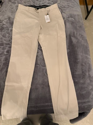 Ljusbeige kostymbyxor från Bläck - Snygga ljusbeige kostymbyxor från Bläck med klassisk rak passform och bälteshällor. Byxorna har två fickor fram och två bak, och är gjorda i mjuk bomull. Perfekta för en clean och stilren look.