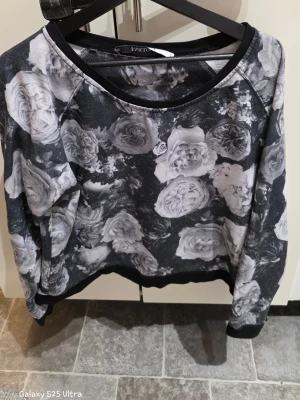 Svart blommig tröja från Fabulous - Säljer en svart tröja från Fabulous i storlek L med stora blommor i grå och vit nyans över hela plagget. Tröjan har rund hals och långa ärmar, perfekt för dig som gillar coola mönster och vill sticka ut lite extra.