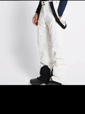 Vita täckbyxor med svarta detaljer - Säljer ett par vita täckbyxor med svarta förstärkningar vid bensluten och svarta hängslen. Byxorna har dragkedjor på sidorna och är tillverkade i ett slitstarkt syntetmaterial, perfekta för vinterns alla äventyr. Vit beigea