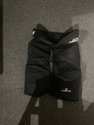 Skyddsbyxor till hockey/bandy jofa. - Ett par svarta hockeybyxor från Jofa. Produkten har vaddering och är tillverkad i slitstarkt syntetmaterial. 