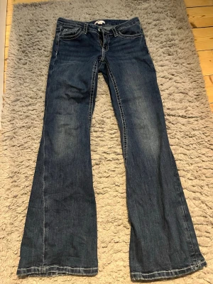 Blå bootcut jeans med broderade fickor - Snygga blå jeans med bootcut passform och kontrastsömmar. Baktill finns två fickor med broderade detaljer i lila och rosa zigzag-mönster samt knappar. Jeansen har klassisk femficksdesign och är tillverkade i jeansmaterial.