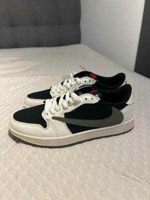 Nike Air Jordan 1 Low Travis Scott - Nike Air Jordan 1 Low Travis Scott med svart och vit ovandel i skinn, olivgrön swoosh och sula. Röda broderade detaljer på hälen och tungan, samt klassisk snörning. Snygg kontrast mellan färgerna och ikoniska loggor gör dessa sneakers till ett måste för sneakerheads.