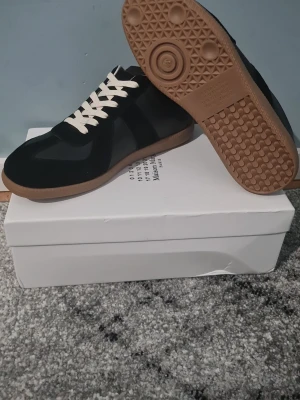 Maison Margiela svarta sneakers - Maison Margiela sneakers i svart mocka och skinn med kontrasterande vita snören och gummisula i beige. Klassisk rund tå och platt sula, stilren design som passar till många outfits. Snygga detaljer och ikonisk Margiela-logga på sulan.