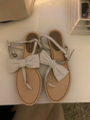 Vita sandaler med rosett - Säljer ett par ljusa sandaler med stora rosetter framtill och smala remmar runt ankeln. De har öppen tå och platt sula! 