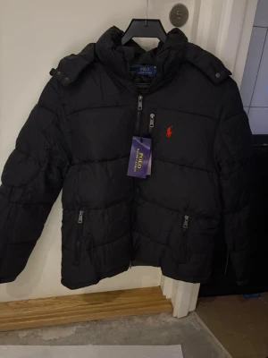 Svart pufferjacka från Polo Ralph Lauren - Säljer en svart pufferjacka från Polo Ralph Lauren med röd logga på bröstet. Jackan har hög krage, huva och flera dragkedjeförsedda fickor. Perfekt för kalla dagar och riktigt snygg till streetwear-stilen. Har i S och m 