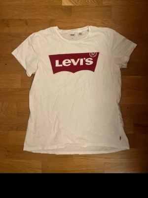 Vit Levi's t-shirt med röd logga - Klassisk vit t-shirt från Levi's i mjuk bomull med den ikoniska röda loggan tryckt på bröstet. T-shirten har rund halsringning, korta ärmar och en liten röd Levi's-tag i sidan. Perfekt basic-plagg för en clean och avslappnad stil.