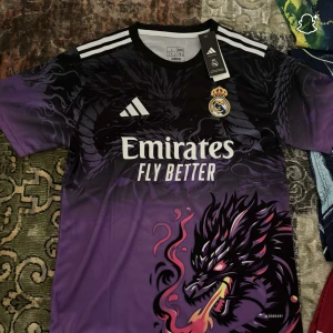 Real Madrid x Adidas lila draktröja - Snygg Real Madrid fotbollströja från Adidas i lila och svart med ett fett drakmotiv på både fram- och baksidan. Kortärmad modell med vita detaljer på axlarna och klubbmärke på bröstet. Materialet är lätt och andas, perfekt för träning eller match.