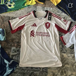 Liverpool vit och röd fotbollströja Adidas - Liverpool FC matchtröja från Adidas i vit med röda detaljer och klubbmärke på bröstet. Kortärmad med svart och röd krage, meshpaneler för ventilation och tryckta sponsorer. Snygg design som sticker ut på planen. Materialet är lätt och andas.