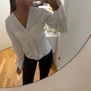 Vit v-ringad skjorta med lång ärm - Säljer en stilren vit skjorta med v-ringning och långa ärmar. Skjortan har figurnära passform och markerad midja som ger en snygg siluett. Perfekt att styla med jeans eller kjol för en clean look.