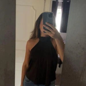 Svart halterneck topp med knyt - Säljer en svart halterneck topp med snygg knyt i nacken och öppen rygg. Toppen är ärmlös och har en loose passform som faller snyggt över kroppen. Perfekt att matcha med jeans eller kjol för en cool look. Perfekt festlig topp samt väldigt elegant, använd en gång.