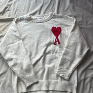 Vit stickad tröja från Ami Paris - Vit stickad tröja från Ami Paris med röd hjärtlogga och bokstaven A på bröstet. Tröjan har rund hals, ribbade muddar och är långärmad. Använd en gång.