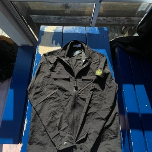 Svart overshirt från Stone Island L - Svart overshirt från Stone Island med klassisk logga på ärmen. Jackan har dragkedja framtill, två bröstfickor och en stilren krage. Tillverkad i lätt syntetmaterial som ger en clean och modern look. Perfekt för lager-på-lager och streetwear-vibe.