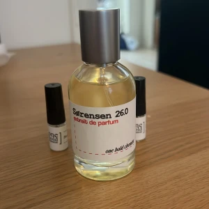 Sørensen 26.0, 50 ml  - Unik parfymflaska från Sørensen, modell 26.0 Extrait de Parfum. Flaskan är rund i genomskinligt glas med silverfärgad kork och vit etikett med svart och röd text. Innehållet är ljust gult. Två mindre provflaskor med svart kork ingår.
