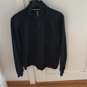 Svart half zip tröja Tommy Hilfiger - Svart långärmad tröja från Tommy Hilfiger med broderad logga framtill. Tröjan har half zip-dragkedja vid halsen och är gjord i en mjuk ekologisk bomullsblandning. Klassisk och stilren design med ribbade muddar och hög krage.
