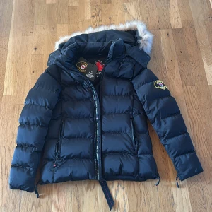Svart pufferjacka från Zavetti Canada - Säljer en svart pufferjacka från Zavetti Canada med huva och fuskpäls. Jackan har broderad logga på ärmen, dragkedjor på fickorna och detaljer med märkesband. Den är helt ny, använd en gång. Gick inte att skicka tillbaka det är därför jag säljer den.