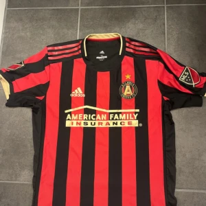 Atlanta United FC matchtröja Adidas  - Snygg Atlanta United FC matchtröja från Adidas med klassiska röda och svarta ränder, guldiga detaljer och klubbmärke på bröstet. Kortärmad modell i lätt och ventilerande ClimaCool-material. MLS-märke på ärmen och tryckt sponsorlogga framtill.