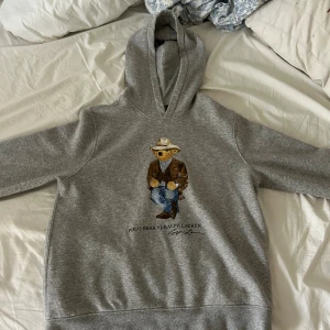 Grå Polo Bear hoodie från Ralph Lauren - Snygg grå hoodie från Polo Ralph Lauren med ikoniska Polo Bear-trycket på bröstet. Björnen är klädd i cowboy-outfit med hatt, mockajacka och jeans. Tröjan har huva och är tillverkad i mjuk bomullsblandning. Perfekt för dig som gillar streetwear med en twist.