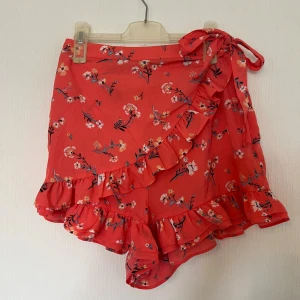 Röda blommiga volangshorts Bik Bok - Snygga röda shorts från Bik Bok med blommigt mönster, volangkant och omlott-design. De har en söt knytning i midjan och är gjorda i mjuk polyester. Perfekta för sommarens alla häng och ger en riktigt fräsch vibe. Endast använda få gånger!