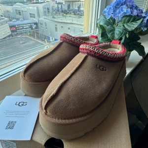 Bruna UGG Tazz boots med broderad kant - Mysiga bruna boots från UGG med fluffigt foder och platt sula. Skorna har en snygg broderad kant i rött och beige samt en rejäl platåsula. Perfekta för kalla dagar när du vill hålla fötterna varma och stiliga.