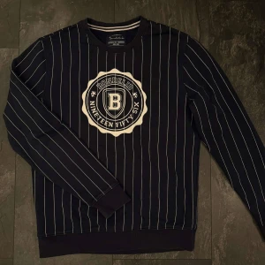 Randig sweatshirt från Bondelid - Snygg marinblå sweatshirt från Bondelid med vita ränder och stort broderat emblem på bröstet. Tröjan har rund hals, långa ärmar och ribbade muddar. Perfekt för dig som gillar klassisk och sportig stil.