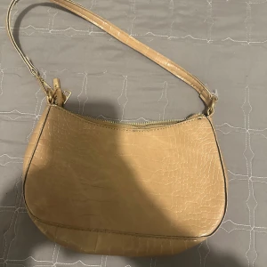 Beige axelväska i skinnimitation - Snygg beige axelväska med croco-mönstrad yta och guldfärgade detaljer. Väskan har en smidig dragkedja upptill och en stilren, rundad form. Perfekt för dig som vill ha en trendig och enkel väska till vardagslooken.