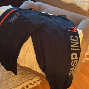Svarta träningsbyxor från Gasp L - Svarta träningsbyxor från Gasp i storlek L med orange detaljer och tryck längs benet. Byxorna har elastisk midja med snörning och är gjorda i ett ventilerande syntetmaterial som passar perfekt för gymmet eller annan träning. helt nya! lappen kvar nypris 800-900kr  mitt pris 600
