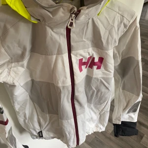 Vit vindjacka Helly Hansen med huva - Snygg vit vindjacka från Helly Hansen med neongul huva och rosa HH-logga på bröstet. Jackan har hel dragkedja framtill, reflexdetaljer och justerbar huva. Perfekt för blåsiga dagar och outdoor-äventyr. Materialet är syntet och jackan har långa ärmar.
