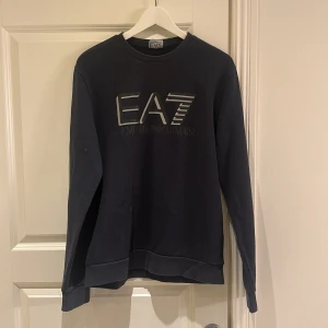Svart EA7 sweatshirt från Emporio Armani - Svart sweatshirt från EA7 Emporio Armani med stor logga framtill. Tröjan har rund hals, långa ärmar och är tillverkad i mjuk bomull. Perfekt för dig som gillar stilrena och exklusiva plagg med streetkänsla.