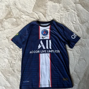 PSG Neymar Jr matchtröja Nike M - Snygg PSG fotbollströja med Neymar Jr och nummer 10 på ryggen. Tröjan är marinblå med vita och röda detaljer, har korta ärmar och är tillverkad i lätt Dri-Fit material. Framsidan har PSG-logga, sponsortryck och klassiska ränder. Perfekt för dig som älskar fotboll och Paris Saint-Germain.