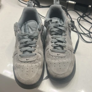 Nike Air Force 1 grå mocka sneakers - Nike Air Force 1 sneakers i grå mocka med svart sula och grå snörning. Swoosh-loggan på sidan har ett unikt mönster i vitt och grått. Klassisk perforerad tå och låg profil. Perfekta för dig som gillar stilrena och coola sneakers med en twist.