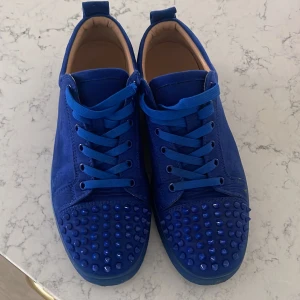 Blå sneakers med nitar och röd sula - Snygga blå sneakers i mocka från Christian Louboutin med ikonisk röd sula. Skorna har snörning och coola nitar över tån för en edgy vibe. Insidan är beige och de har en klassisk låg profil. Perfekta för dig som vill sticka ut med din stil. Man får med låda!
