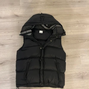 Svart dunväst från Burberry - Snygg svart puffer vinter jacka  från Burberry London England med avtagbar armar och dragkedja framtill. Västen har två fickor med dragkedja och ett klassiskt rutigt foder i huvan gjort i ull. Först till kvarn