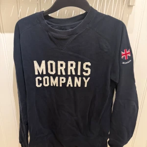 Mörkblå sweatshirt Morris Company - Mörkblå sweatshirt från Morris Company med broderad logga framtill och brittisk flagga på ärmen. Klassisk rund halsringning och långa ärmar. Perfekt för dig som gillar stilren och sportig look.