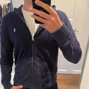 Mörkblå hoodie från Polo Ralph Lauren - Mörkblå hoodie från Polo Ralph Lauren med dragkedja, huva med snörning och klassisk broderad logga på bröstet. Tröjan har känguruficka framtill och är tillverkad i mjuk bomull. Tröjans storlek lyder XL men har krympt sedan innan jag köpte (2nd hand) och passar S-M 