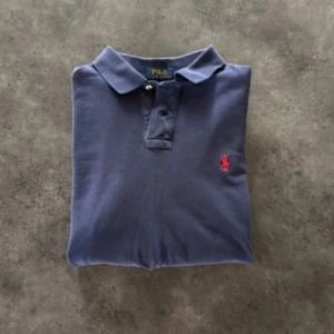 Mörkblå långärmad pikétröja Polo Ralph Lauren - Säljer en klassisk mörkblå långärmad pikétröja från Polo Ralph Lauren med röd broderad logga på bröstet. Tröjan har krage och knäppning framtill, perfekt till jeans eller chinos. Tillverkad i mjuk bomull för en skön känsla.