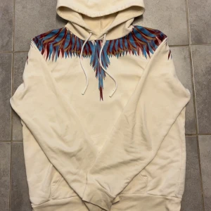Marcelo Burlon hoodie - Säljer en gräddvit hoodie från Marcelo Burlon County of Milan med ikoniskt färgglatt fjädermönster i blått, rött och lila över axlar och bröst. Tröjan har huva med snörning och stor magficka. Perfekt statement-plagg för dig som vill sticka ut. Storlek M. Modellen är 185 och väger ca 70 kg. Hör gärna av dig om det finns frågor☺️
