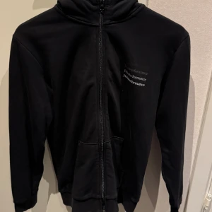 Svart hoodie från Peak Performance - Svart hoodie från Peak Performance med dragkedja framtill och huva. Klassisk design med logga på bröstet och två fickor. Bra skick!