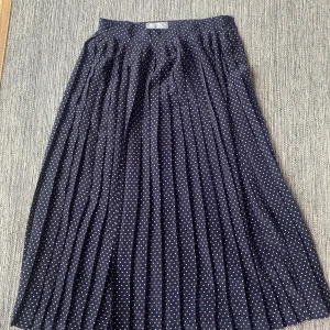 Mörkblå plisserad kjol med hjärtan - Supersöt mörkblå långkjol från Prêt à Porter med vita hjärtan över hela tyget. Kjolen är plisserad och har en klassisk midja. Perfekt för dig som gillar romantiska detaljer och vill sticka ut med ett gulligt mönster.