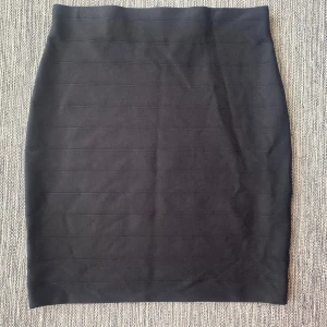 Svart tight kjol i stretchigt tyg - En svart, figurnära kortkjol med diskreta horisontella sömmar. Kjolen har en enkel och stilren design, perfekt för dig som gillar minimalistiskt mode. Tillverkad i ett stretchigt material som sitter snyggt och bekvämt.