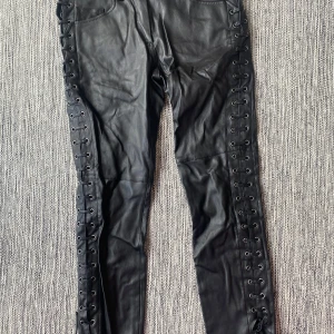 Svarta skinnbyxor med snörning - Snygga svarta skinnbyxor med snörning längs båda sidorna och coola fickdetaljer bak. Byxorna har en slim passform och är gjorda i skinn. Perfekta för dig som vill sticka ut med edgy stil och detaljer som verkligen syns.