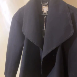 Svart kappa med bred krage - Snygg svart kappa med bred, markerad krage och rak passform. Jackan har lång ärm och är tillverkad i ett tjockare tyg som ger en stilren look. Perfekt för dig som gillar minimalistisk stil och vill ha en jacka som sticker ut med sin coola krage.